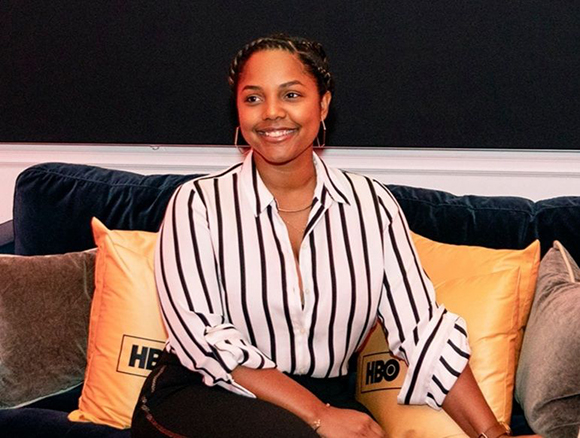 A smiling woman with braided hair wearing a black and white striped shirt sits on a blue sofa with yellow pillows.