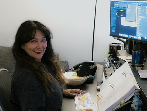A woman with long dark hair sits at a desk with a computer monitor, keyboard, and various office supplies.