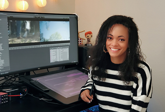 A smiling woman with curly dark hair wearing a striped sweater sits at a desk with a large monitor.