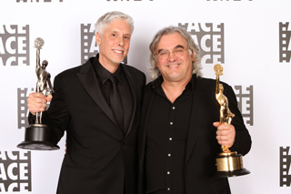 Two men in black suits smiling and holding gold trophies against a white patterned backdrop.