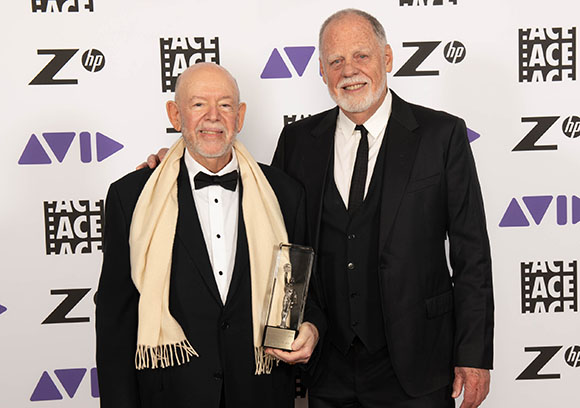 Two smiling men in formal black attire pose together while holding a tall glass trophy.
