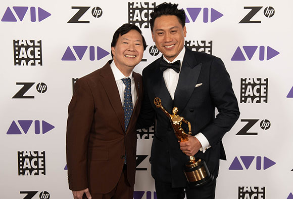 Two men smiling; one in a brown suit and one in a black tuxedo holding a gold trophy.