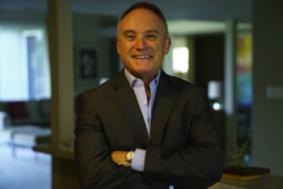 A smiling man in a dark suit and light blue shirt stands with his arms crossed in a blurred indoor setting.