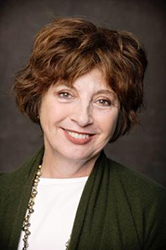 A smiling woman with short reddish-brown hair wearing a white top, green cardigan, and a beaded necklace.