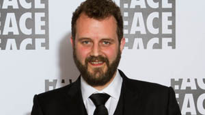 A man with a brown beard and hair wearing a black suit, white shirt, and black tie.