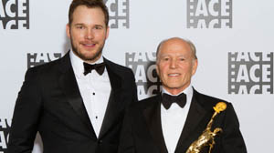 Two men in black tuxedos and bowties posing in front of a white backdrop with logos.