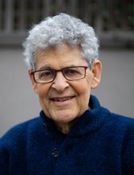 An elderly person with curly grey hair and glasses smiling at the camera.