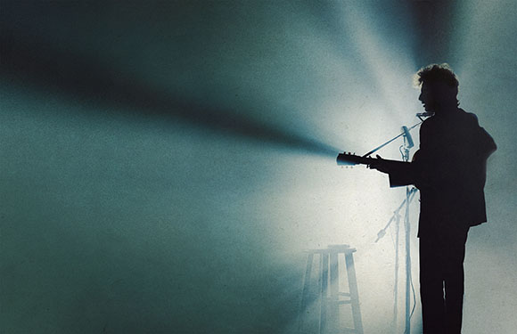 Silhouette of a person playing a guitar under a bright beam of light on a dark stage.