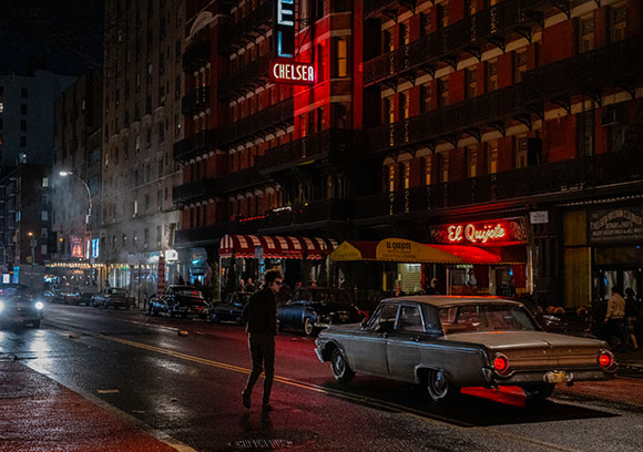A person walks across a wet city street at night past the illuminated Chelsea Hotel and vintage cars.