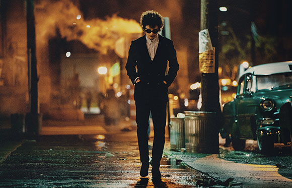 A man in a dark suit and sunglasses walks down a wet city street at night.