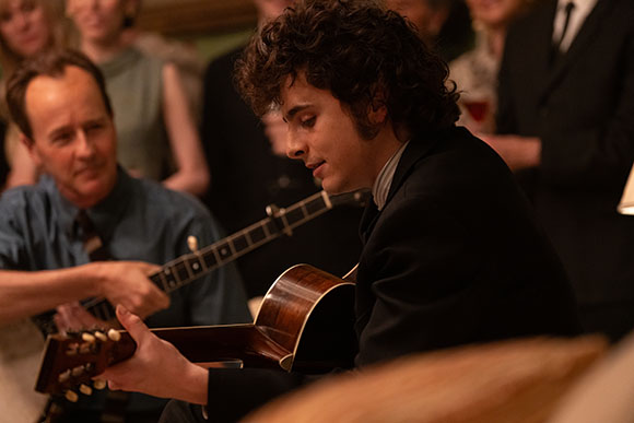 A young man with curly hair plays an acoustic guitar while a man in a blue shirt watches in the background.