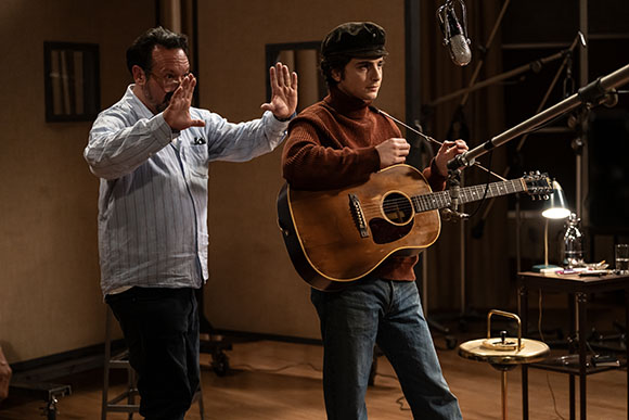 A man in a striped shirt gestures with open palms toward a man holding an acoustic guitar.
