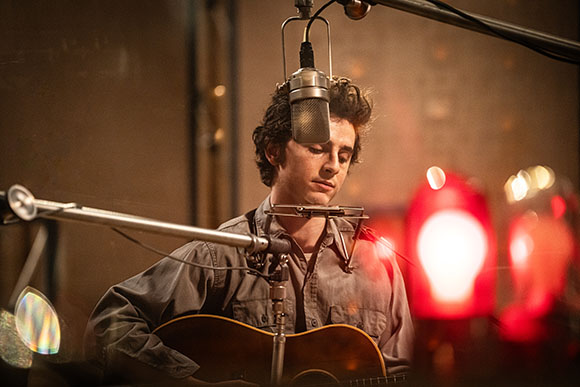 A man with curly hair plays an acoustic guitar in a studio with microphones and red lights.