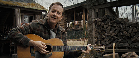 A man in a plaid shirt playing an acoustic guitar in front of a rustic wooden cabin.