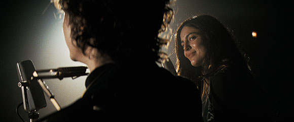 A person in silhouette faces a smiling woman in a dimly lit setting with a microphone nearby.