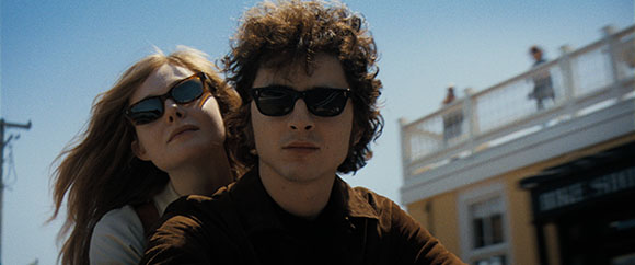 A man and a woman with sunglasses look forward against a bright blue sky and a building balcony.