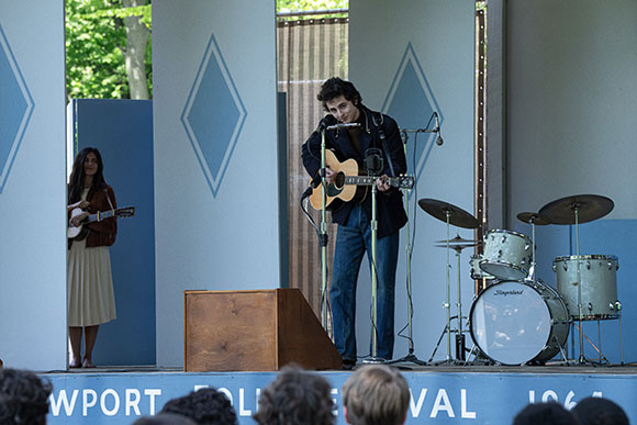 A man plays an acoustic guitar on a stage next to a drum kit and a woman standing in a doorway.