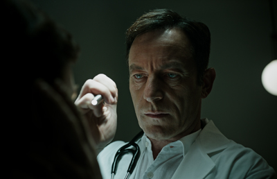 A male doctor in a white coat looks intently at a patient while holding a medical tool in a dimly lit room.