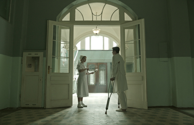 A nurse in white holds a clipboard while a man on crutches stands in a large arched doorway.