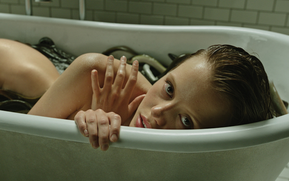 A woman with wet hair reclines in a white bathtub filled with dark liquid and tangled black cables.