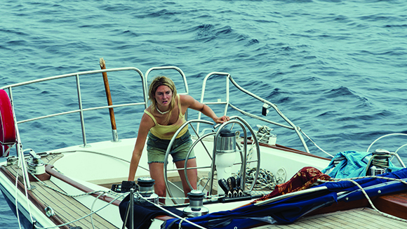 A woman in a yellow tank top and denim shorts stands on the deck of a sailboat on the ocean.
