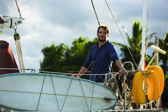 A man in a blue shirt stands on a sailboat next to a white surfboard and yellow buoys.
