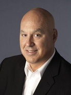 A bald man wearing a black suit jacket and a white collared shirt smiling against a gray background.