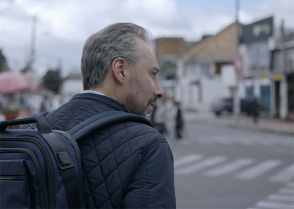 A man with graying hair wearing a quilted navy jacket and a backpack looks back on a city street.