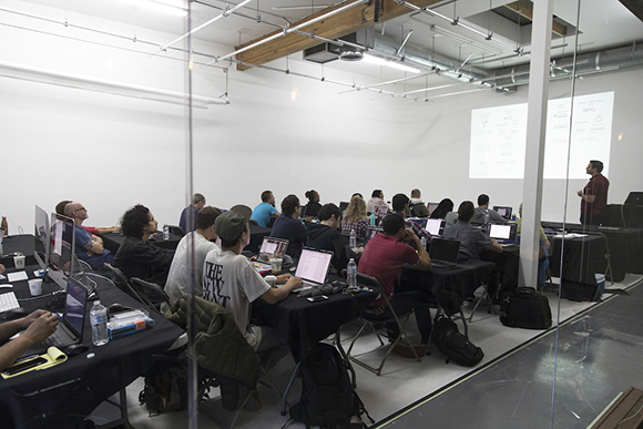 A classroom setting where people sit at long tables with laptops, facing a presenter and a projection screen.