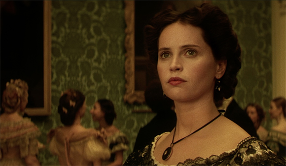 A woman with dark hair in an updo wearing a dark necklace and patterned dress in a vintage room.