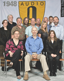 Seven people of various ages and genders posing together for a group portrait against a light background.