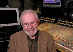 A smiling man with glasses and a beard sitting in front of a large mixing console in a studio.