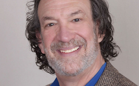 A smiling man with wavy dark hair and a graying beard wearing a blue shirt and brown jacket.