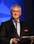 A man in a black suit and light blue shirt speaking into a microphone while holding papers.