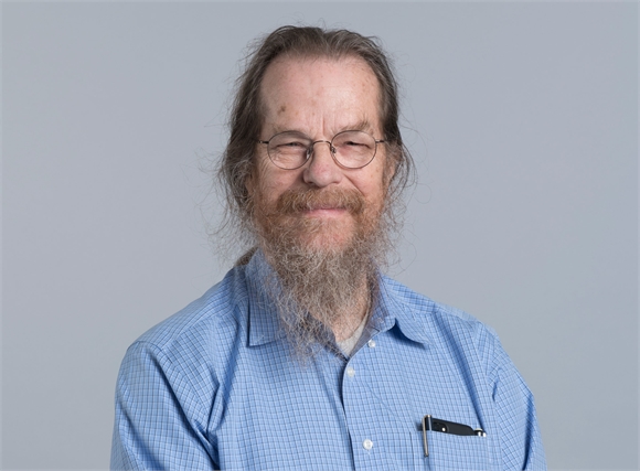 A man with long brown hair, a graying beard, and glasses wearing a blue checkered shirt.