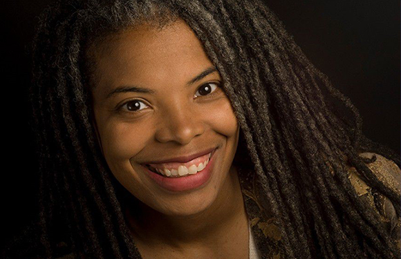 A smiling woman with long, dark dreadlocks against a black background.