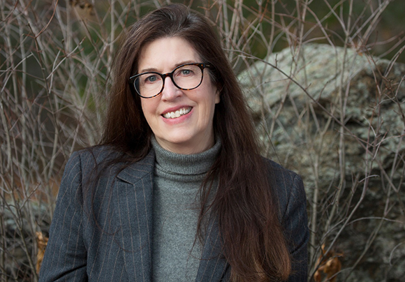 A smiling woman with long dark hair and glasses wearing a grey turtleneck and a pinstripe blazer.