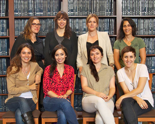 Seven women posing in two rows in front of a tall bookshelf filled with dark books.