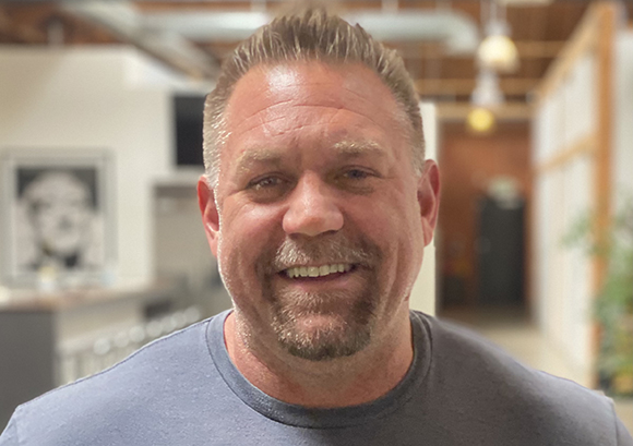 A smiling man with short brown hair and a goatee wearing a grey t-shirt.