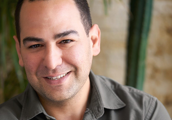 A smiling man with short dark hair and light stubble looking toward the camera.
