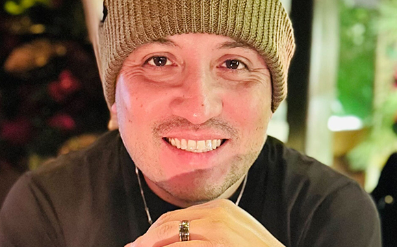 A smiling man wearing a brown knit beanie and a black shirt.