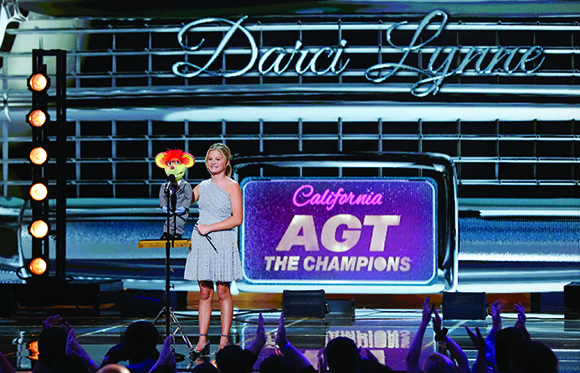 A young girl in a silver dress stands next to a puppet on a stage with a large sign reading AGT The Champions.