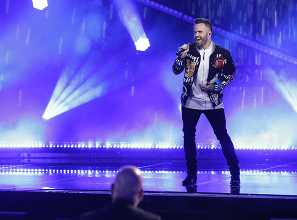 A man in a patterned jacket sings into a microphone on a stage with blue lights and fog.