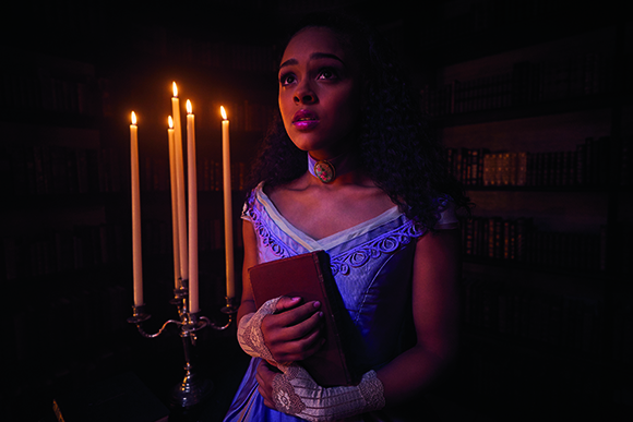 A young woman looks upward while holding a book in a dimly lit room filled with candles and bookshelves.