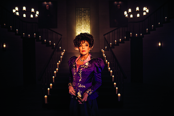 A woman in a purple ornate gown stands centered before a dark, candlelit grand staircase.