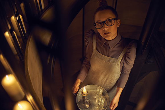 High-angle view of a person in glasses and an apron holding a tray with lit candles in a dim setting.