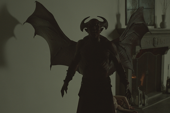 A dark, horned, winged demonic figure stands in a room near a fireplace.