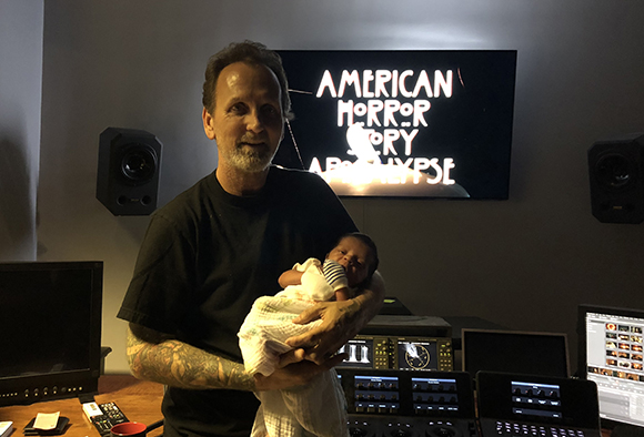 A man with tattoos holds a baby in front of a glowing American Horror Story Apocalypse sign in a studio.