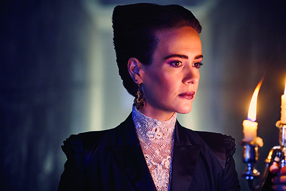 A woman with dark hair in an updo wearing a lace collar and dark blazer, looking right near lit candles.