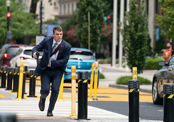 A man in a blue suit and tie runs across a city crosswalk between yellow and black bollards.